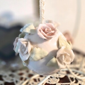 May include: A ceramic ornament, spherical in shape, adorned with delicate, pastel-colored roses and leaves. The ornament is a soft pink hue, with the roses in shades of pink and cream. A lace ribbon is attached for hanging.