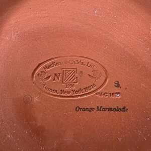 May include: Close-up of a terracotta-colored ceramic plate with the embossed text "Mackenzie-Childs, Ltd" and "Orange Marmalade". The plate has a circular emblem with the year 1996 and the location "Aurora, New York 13026".