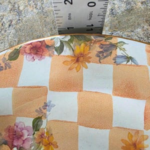 May include: A decorative plate with a checkered pattern in orange and white, adorned with floral designs in shades of pink, yellow, and blue. The plate has a gold rim and is partially measured with a ruler.
