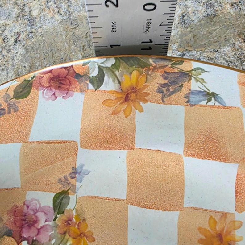 May include: A decorative plate with a checkered pattern in orange and white, adorned with floral designs in shades of pink, yellow, and blue. The plate has a gold rim and is partially measured with a ruler.