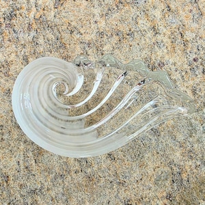 May include: A clear glass decorative piece with a spiral design. The glass has a wave-like shape with a swirling pattern on one end. The piece is transparent and sits on a textured surface.