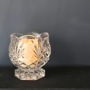 May include: A clear, cut-glass candle holder with a lit candle inside. The candle holder has a tulip shape with intricate leaf and diamond patterns. The base is a hexagonal shape. The candle flame glows warmly.
