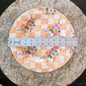 May include: A decorative plate with a checkered orange and white pattern, adorned with floral arrangements. The plate, approximately 12 inches in diameter, features a gold rim and is set against a neutral background.