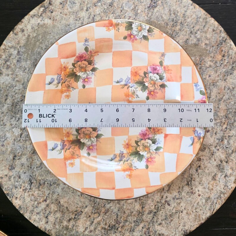 May include: A decorative plate with a checkered orange and white pattern, adorned with floral arrangements. The plate, approximately 12 inches in diameter, features a gold rim and is set against a neutral background.