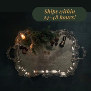 May include: A rectangular silver-toned serving tray with ornate, scalloped edges and two side handles. The tray is decorated with a lit candle in a holder, a sprig of green pine needles, several small brown nuts, and a small metal bell.