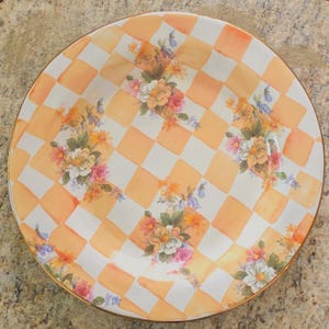 May include: A round, decorative plate with a gold rim. The plate features a checkered pattern in orange and white, with floral bouquets in the center. The flowers are in shades of pink, orange, and white.