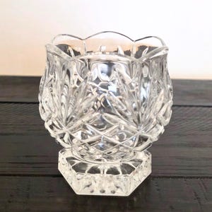 May include: A clear, cut-glass votive candle holder with a scalloped rim. The sides feature a diamond and leaf pattern, and it sits on a hexagonal base. The candle holder is designed to hold a small candle.