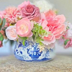May include: A white ceramic bowl featuring a blue floral pattern and a central urn motif, filled with an arrangement of artificial flowers in shades of pink, peach, and cream, accented with green leaves and small sprigs of foliage.