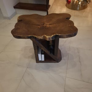 May include: A wooden side table with a live edge top and a unique, angled shelf underneath. The table is made of dark brown wood and has a natural, rustic look.