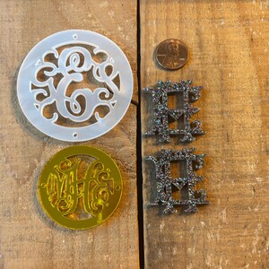 Könnte beinhalten: Vier Dekorationsstücke für ein Zuhause, darunter eine weiße Kunststoffschablone eines Monogramms, ein goldenes Spiegelmonogramm und zwei schwarze Glitzerbuchstaben, die "HOME" buchstabieren.