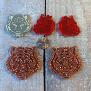 May include: Four decorative ornaments featuring animal faces. Two are red and two are gold. One gold ornament is a tiger face, and the other is a tiger face with glitter. One red ornament is a bulldog face, and the other is a bulldog face with glitter.