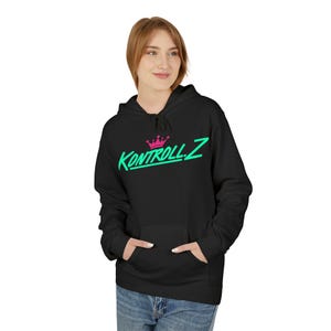 Peut inclure: Sweat à capuche noir avec une poche avant. Le sweat-shirt affiche le texte "Kontroll.Z" en vert fluo avec une couronne rose au-dessus. Le modèle porte un jean bleu.