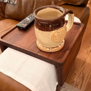 May include: A rectangular wooden armrest tray with a dark brown finish, featuring a ceramic mug with a two-tone brown and cream glaze and a Big Canoe label, a black remote control, and a round wooden coaster with a white leaf pattern design.
