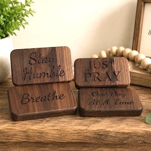 May include: Four rectangular dark brown wooden blocks with rounded corners. The top left block is engraved with Stay Humble, the bottom left with Breathe, the top right with JUST PRAY, and the bottom right with One Day At A Time.