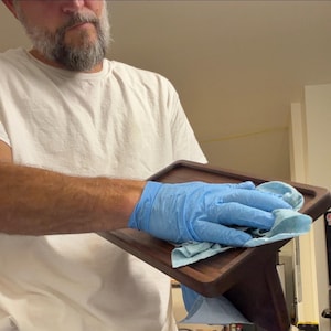 May include: A rectangular dark brown wooden tray with rounded corners and raised edges. The tray has a smooth, polished finish and is being wiped down with a light blue cloth by a hand wearing a blue nitrile glove.