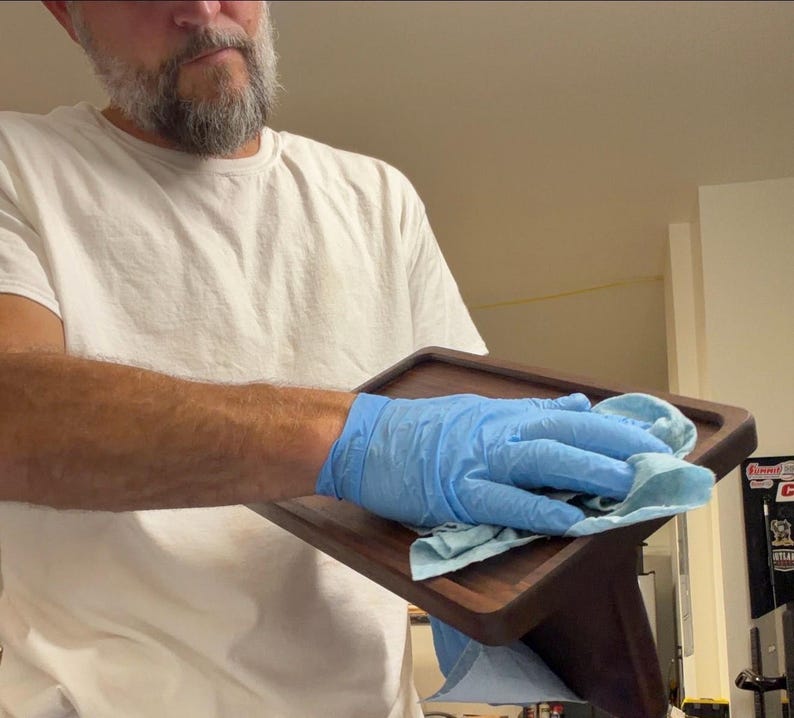 May include: A rectangular dark brown wooden tray with rounded corners and raised edges. The tray has a smooth, polished finish and is being wiped down with a light blue cloth by a hand wearing a blue nitrile glove.