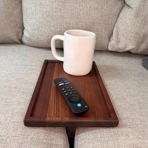 May include: A rectangular dark wood sofa arm tray with a smooth finish, featuring a white ceramic mug and a black remote control with various buttons placed on its surface, designed to rest securely over the arm of a couch.