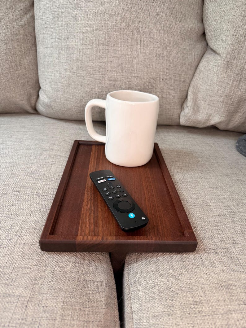 May include: A rectangular dark wood sofa arm tray with a smooth finish, featuring a white ceramic mug and a black remote control with various buttons placed on its surface, designed to rest securely over the arm of a couch.