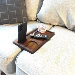 May include: A dark brown wooden sofa arm tray with a built-in phone holder, a circular cutout, and a rectangular surface. A black phone, a spoon, a remote control, and a yogurt cup are on the tray. The tray is on a light-colored sofa.