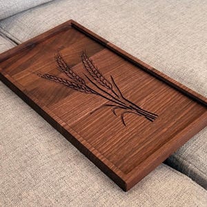 May include: A rectangular dark brown wooden serving tray featuring a laser-engraved design of three wheat stalks across the center. The tray has a smooth finish with visible wood grain patterns and a raised border edge around the entire perimeter.