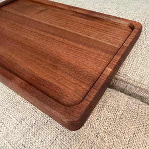May include: A rectangular wooden serving tray with a smooth, dark brown finish and a recessed center area. The tray features rounded corners and a raised border edge, showcasing the natural wood grain pattern across its flat surface.