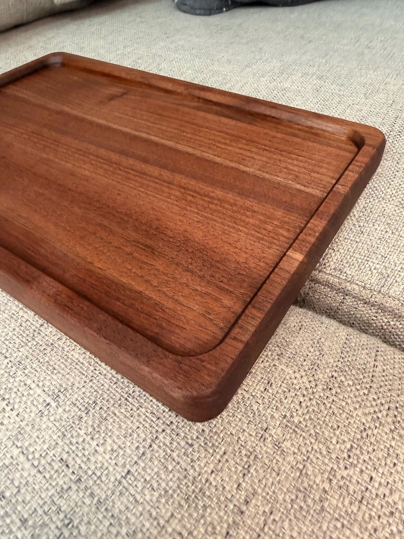 May include: A rectangular wooden serving tray with a smooth, dark brown finish and a recessed center area. The tray features rounded corners and a raised border edge, showcasing the natural wood grain pattern across its flat surface.