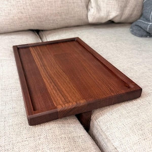 May include: A rectangular dark brown wooden tray with a smooth, flat surface and a raised border edge. The tray features a support piece underneath, designed to rest securely across the arm of a sofa, providing a stable flat surface.