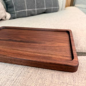 May include: A rectangular dark brown wooden tray with a smooth, polished surface and slightly raised edges. The tray features a visible natural wood grain pattern throughout its flat base and rounded corners, showcasing a deep, rich walnut-toned finish.