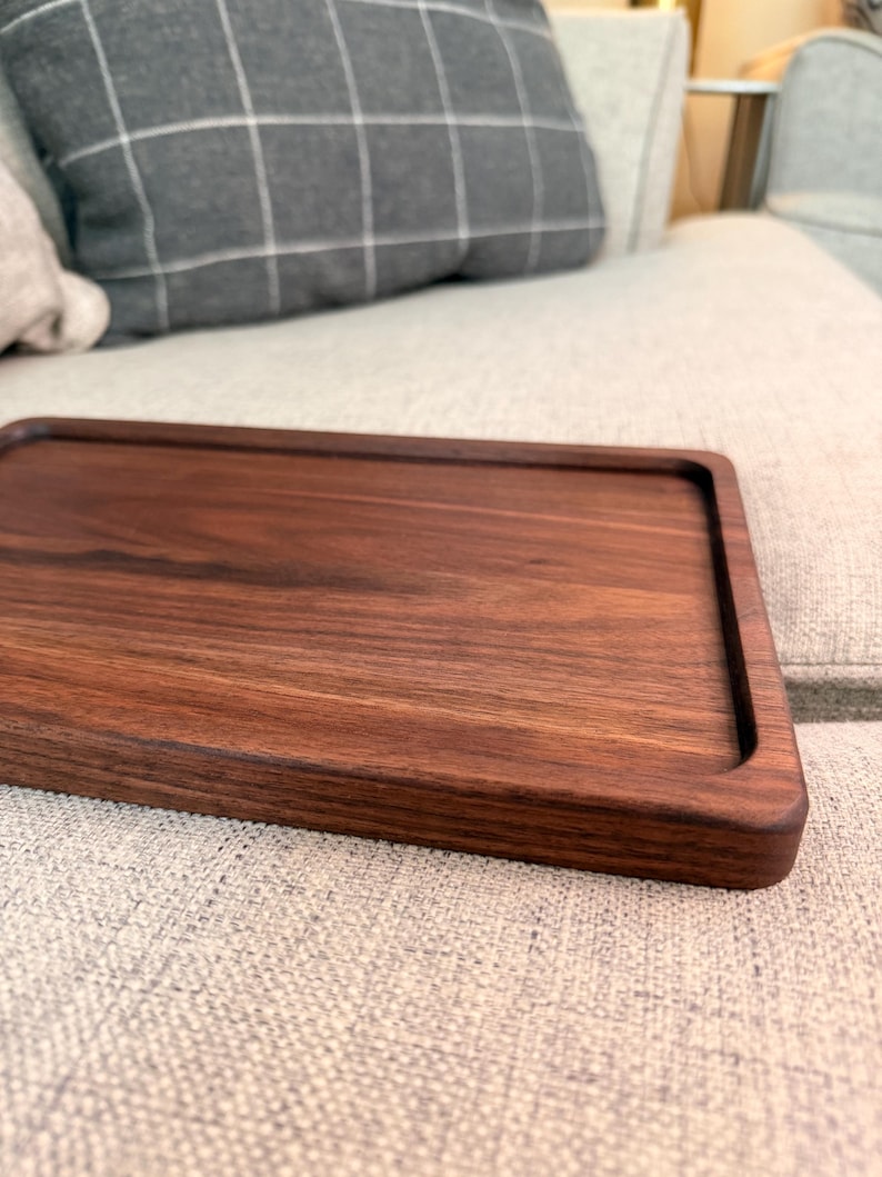 May include: A rectangular dark brown wooden tray with a smooth, polished surface and slightly raised edges. The tray features a visible natural wood grain pattern throughout its flat base and rounded corners, showcasing a deep, rich walnut-toned finish.
