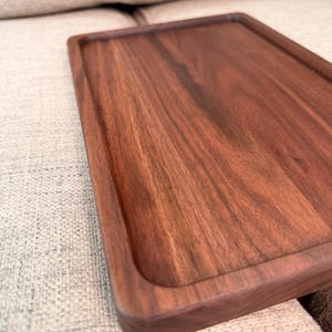 May include: A rectangular wooden tray with a smooth, dark brown finish and visible natural wood grain patterns. The tray features a raised border around the perimeter, creating a recessed surface area with rounded corners and straight edges.