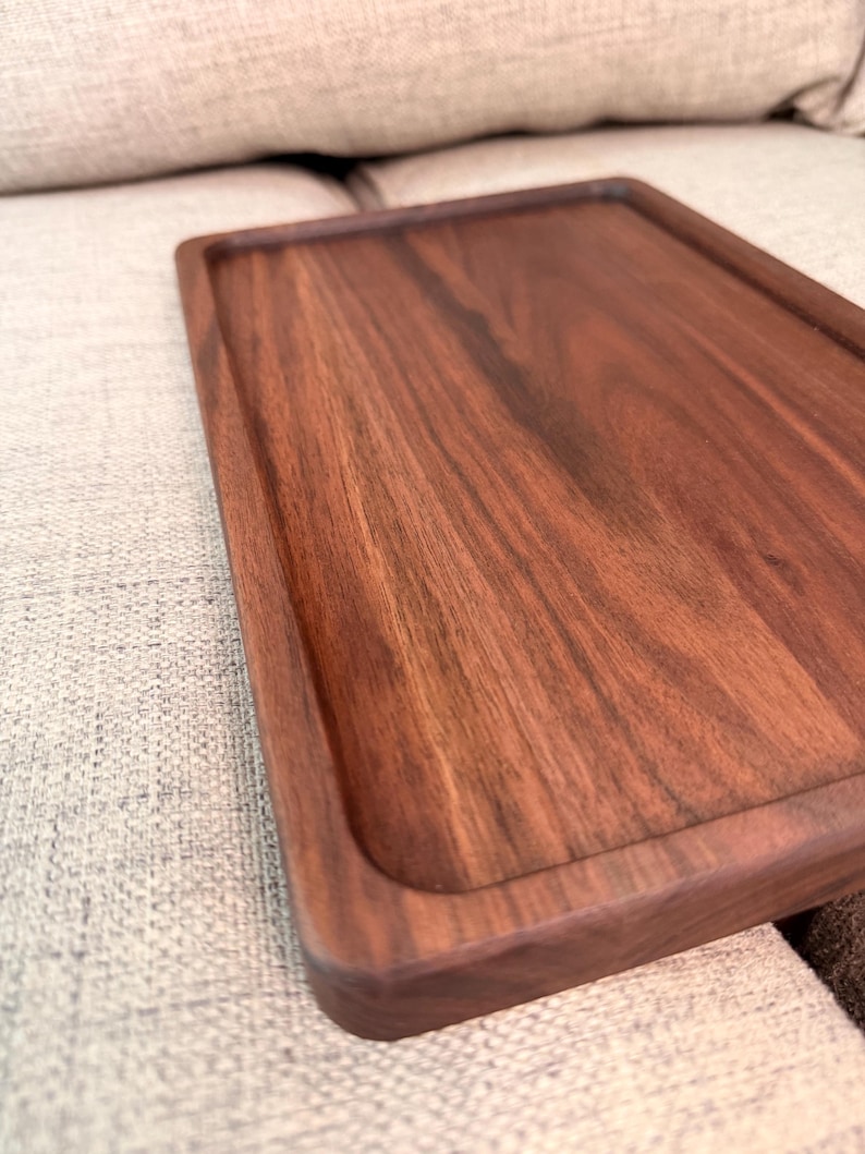 May include: A rectangular wooden tray with a smooth, dark brown finish and visible natural wood grain patterns. The tray features a raised border around the perimeter, creating a recessed surface area with rounded corners and straight edges.