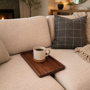 May include: A rectangular dark brown wooden tray rests on a beige fabric sofa. A speckled off-white ceramic mug filled with black coffee sits on a small round wooden coaster placed on the center of the tray.