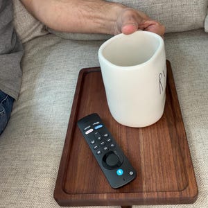 May include: A rectangular dark brown wooden tray resting on a couch, holding a white ceramic mug with partial black lettering and a black television remote control with various buttons and a blue power icon.