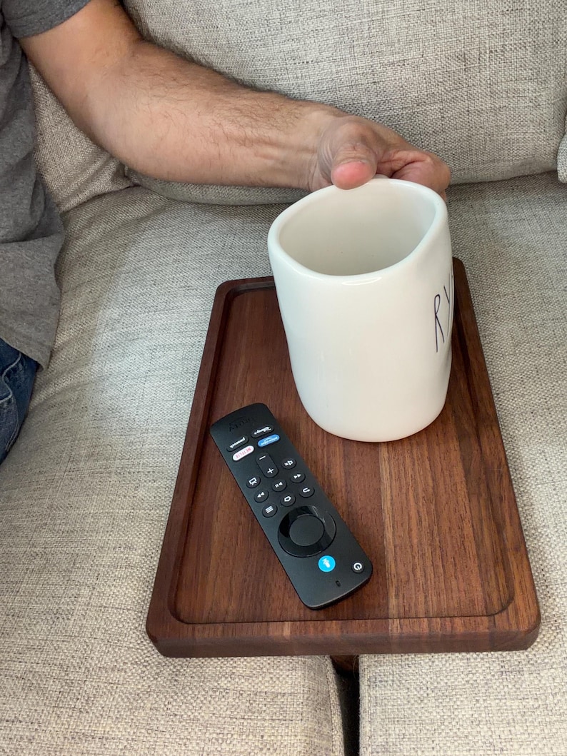 May include: A rectangular dark brown wooden tray resting on a couch, holding a white ceramic mug with partial black lettering and a black television remote control with various buttons and a blue power icon.