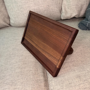 May include: A rectangular dark brown wooden stand with a smooth, polished finish and a recessed center panel. The stand is angled upward, featuring a sturdy base support on the back, showcasing the natural wood grain patterns throughout the surface.