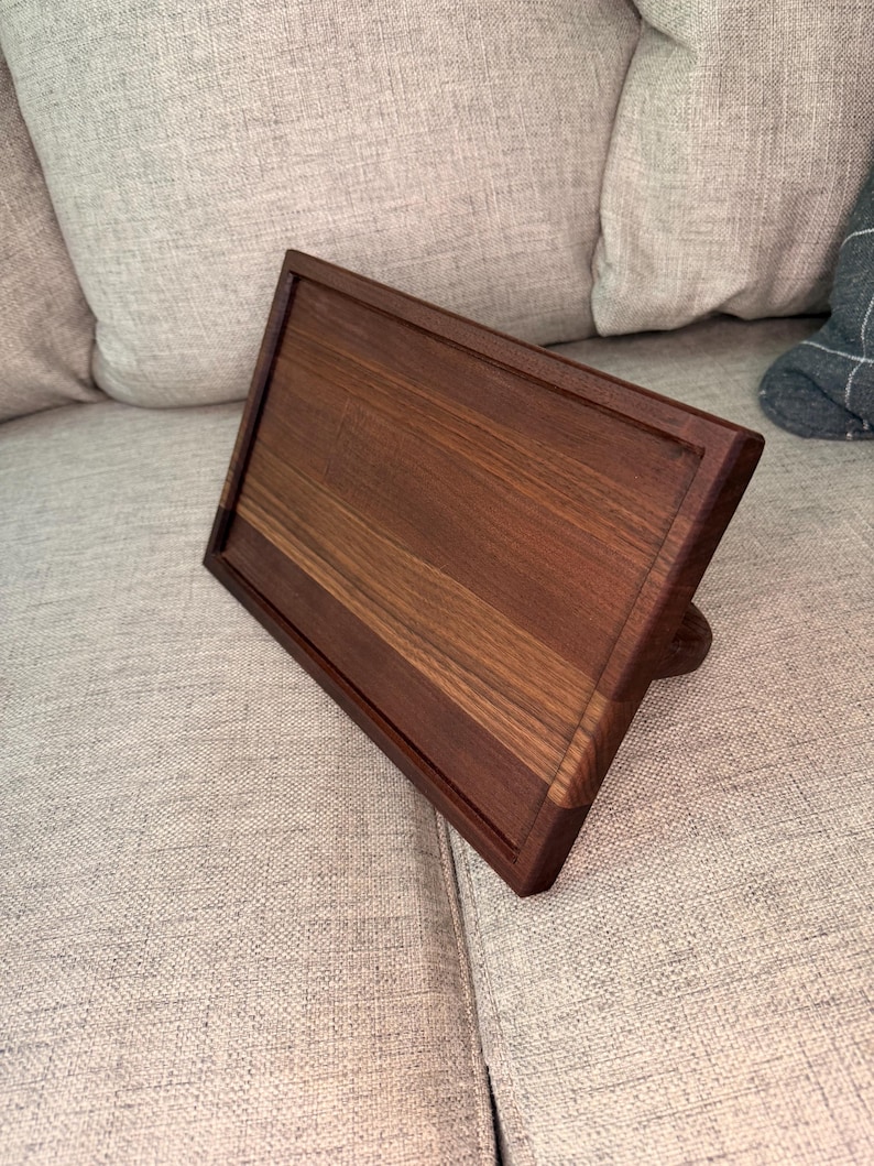 May include: A rectangular dark brown wooden stand with a smooth, polished finish and a recessed center panel. The stand is angled upward, featuring a sturdy base support on the back, showcasing the natural wood grain patterns throughout the surface.
