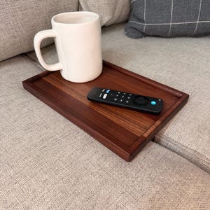 May include: A rectangular dark brown wooden tray with a smooth finish, holding a plain white ceramic mug and a black remote control with various buttons, including a prominent blue circular button, resting on a textured fabric surface.