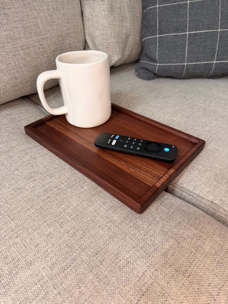 May include: A rectangular dark brown wooden tray with a smooth finish, holding a plain white ceramic mug and a black remote control with various buttons, including a prominent blue circular button, resting on a textured fabric surface.