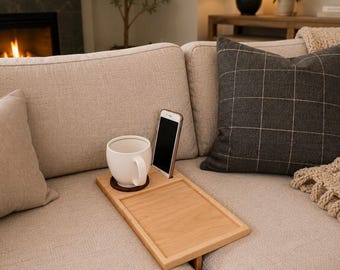 May include: A rectangular light-colored wooden sofa arm tray featuring a dedicated circular cutout for a mug and a narrow slot for a smartphone. The tray rests securely over the arm of a beige couch, holding a white ceramic mug and a phone.