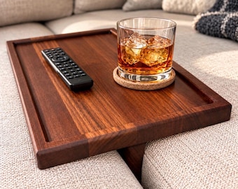May include: A rectangular wooden sofa arm tray with a dark brown finish. The tray holds a black television remote control and a clear glass tumbler filled with an amber liquid and ice cubes, resting on a round cork coaster.