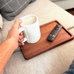 May include: A rectangular wooden tray with a smooth finish, featuring a white ceramic mug and a black television remote control placed on its surface. The tray is designed with a slightly raised edge to keep items secure while resting.