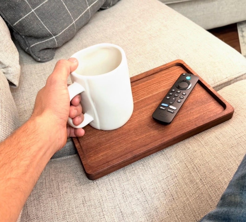 May include: A rectangular wooden tray with a smooth finish, featuring a white ceramic mug and a black television remote control placed on its surface. The tray is designed with a slightly raised edge to keep items secure while resting.