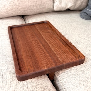 May include: A rectangular, dark brown wooden tray with a raised edge. The tray is placed on a light beige sofa. The wood grain is visible, and the tray appears to be made of a solid piece of wood.