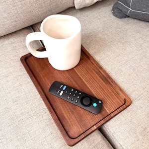 May include: A rectangular dark brown wooden serving tray featuring a smooth finish and raised edges. A large white ceramic mug with a rounded handle and a black television remote control with various buttons are placed on the tray surface.