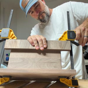 May include: A rectangular piece of dark brown walnut wood is being held in place by two yellow and black plastic bar clamps. The wood has a smooth, natural grain pattern and is positioned horizontally while being secured for assembly.