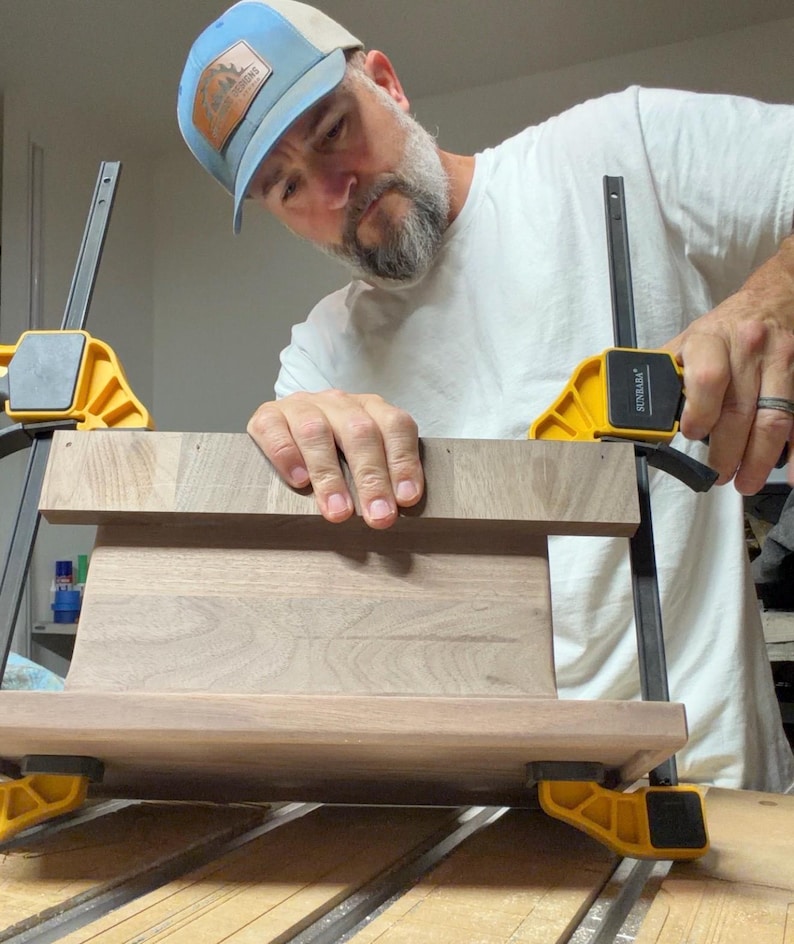 May include: A rectangular piece of dark brown walnut wood is being held in place by two yellow and black plastic bar clamps. The wood has a smooth, natural grain pattern and is positioned horizontally while being secured for assembly.