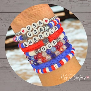 May include: A stack of patriotic bracelets in red, white, and blue. Some bracelets spell out "USA AMERICA" with letter beads. Other bracelets are made of colorful beads and flat, disc-shaped beads. The bracelets are stacked on a wrist.