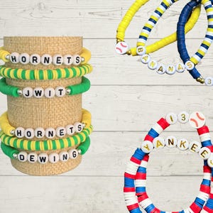 May include: A collection of colorful beaded bracelets. Some bracelets feature team names like "Hornets" and "Yankees" spelled out with white letter beads. Other bracelets have baseball-themed charms and are in team colors like blue, yellow, red, and white.