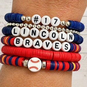 May include: Stack of beaded bracelets in red, blue, and silver. One bracelet features the text "#17", another "LINCOLN BRAVES", and a baseball charm. The bracelets are made of flat, disc-shaped beads.