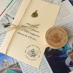 May include: A beige card tied with twine, featuring a sailboat charm and the text "wish you joy...wish every happiness...always be with you." A wooden coaster with "FROM THE LIBRARY OF PAMELA BAKER" is also present.